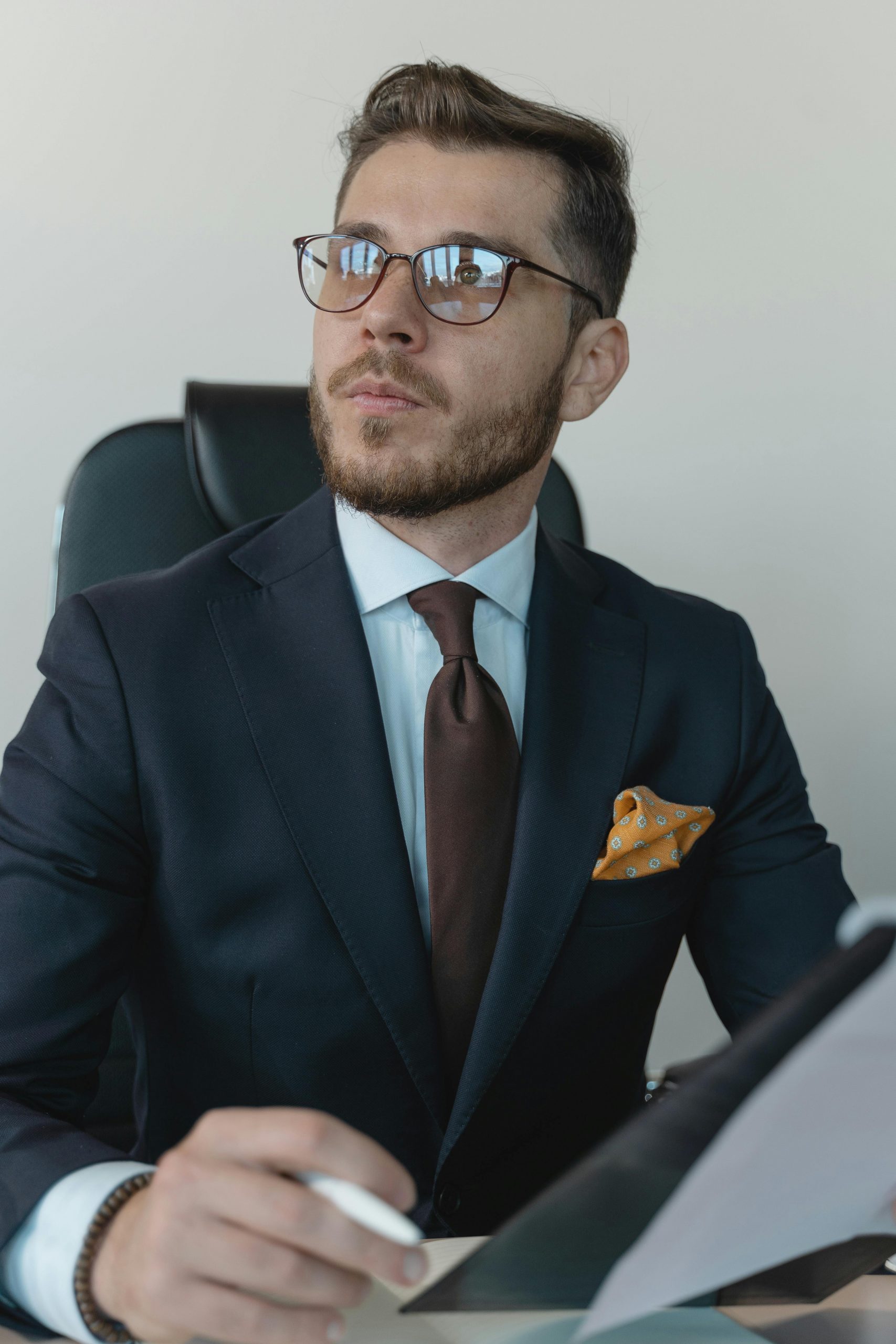 Young professional businessman wearing a suit and glasses, sitting at a desk with documents.